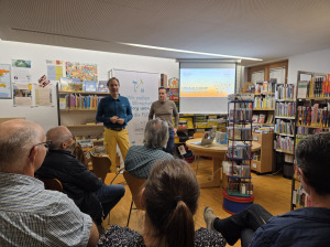 zwei stehende Menschen die sprechen - fünf sitzende Menschen von hinten die zuhören. Vortragssetting