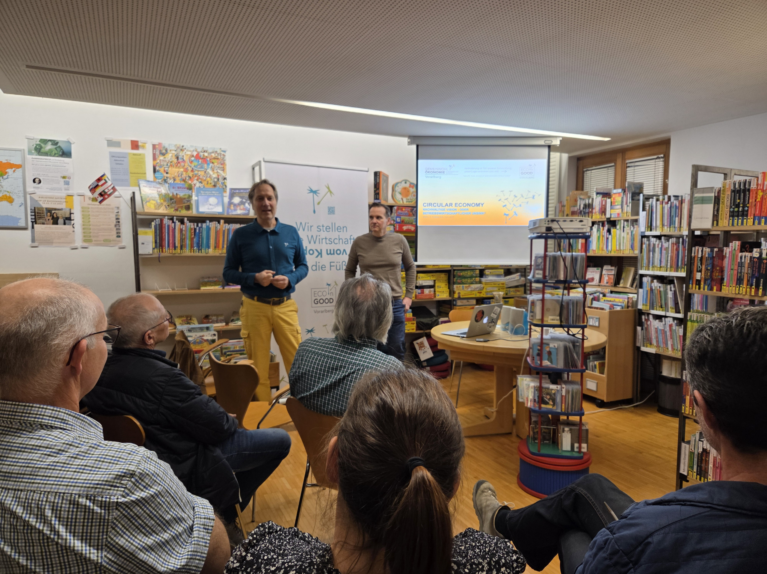 zwei stehende Menschen die sprechen - fünf sitzende Menschen von hinten die zuhören. Vortragssetting